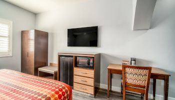 The image shows a hotel room with a bed, a wall-mounted TV, a desk with a chair, a wardrobe, and a microwave on a cabinet.