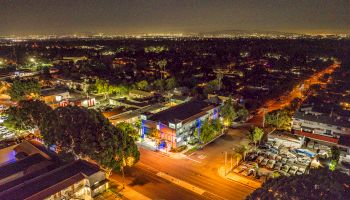 Night aerial view of a cityscape with illuminated streets and buildings, surrounded by trees and distant city lights.