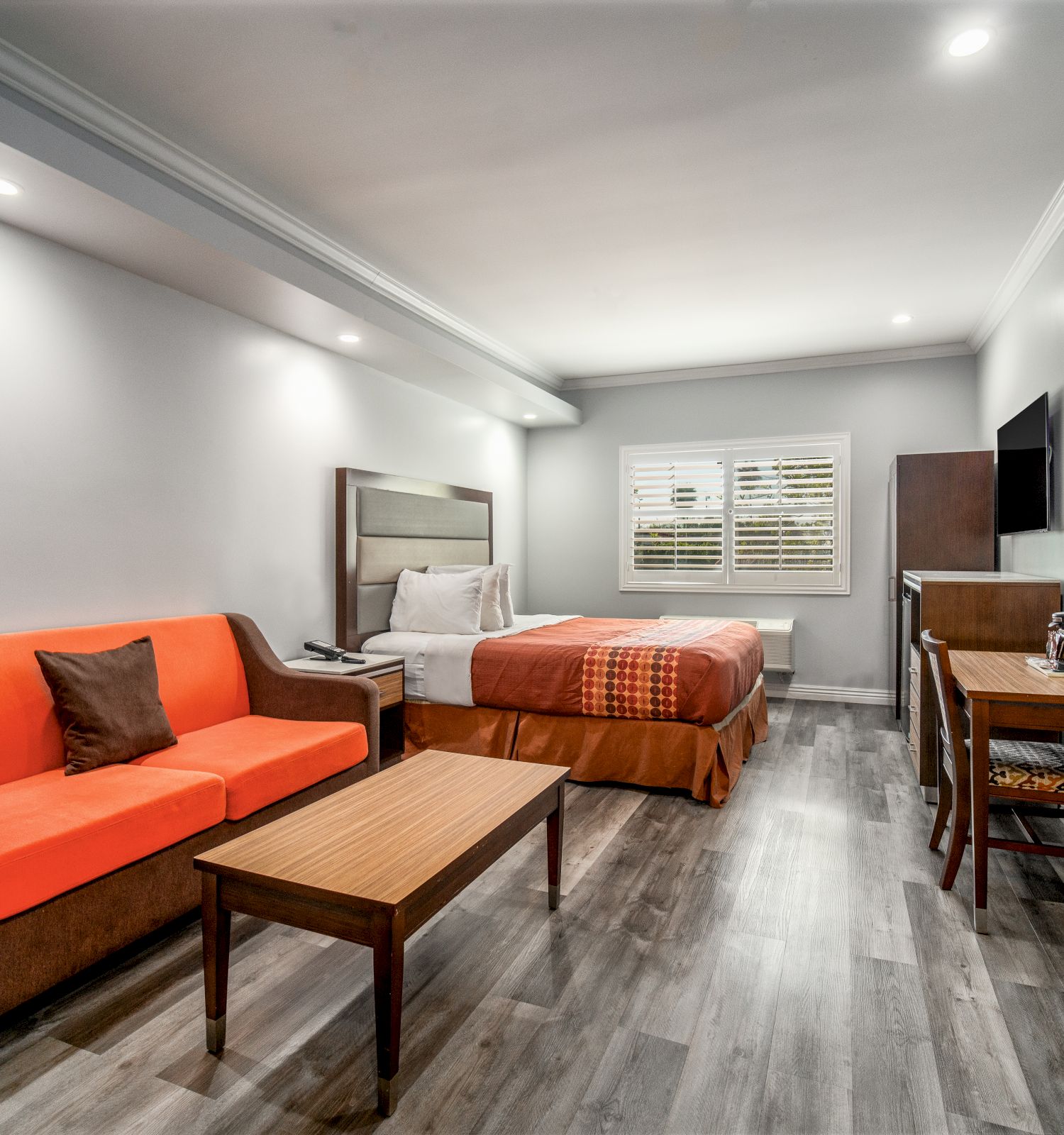 The image shows a modern hotel room with a bed, orange sofa, desk, TV, and wooden flooring, creating a cozy and functional space.