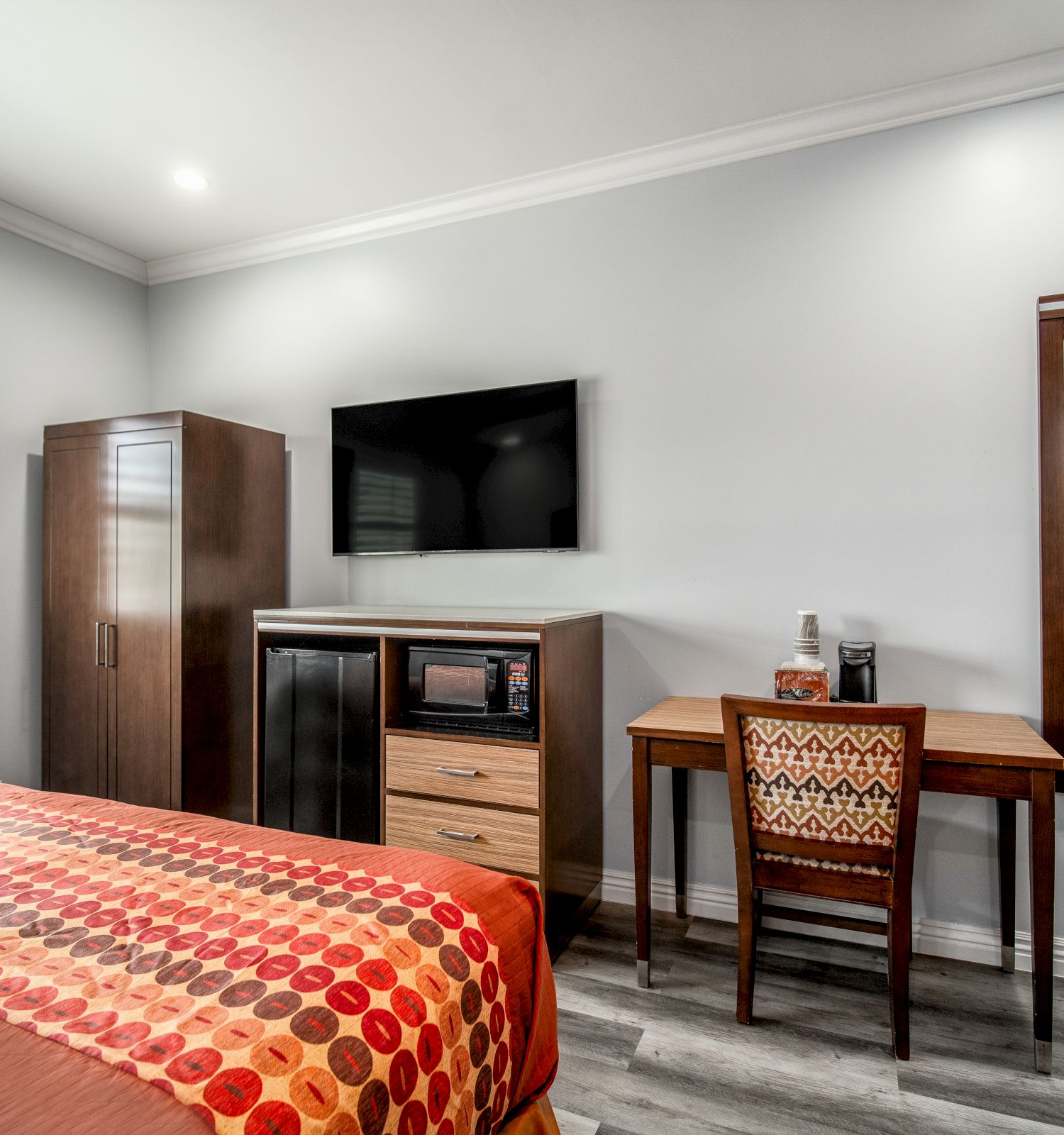 A modern room with a bed, TV, desk, chair, mirror, and cabinet. The decor is simple with wooden furniture and light-colored walls.