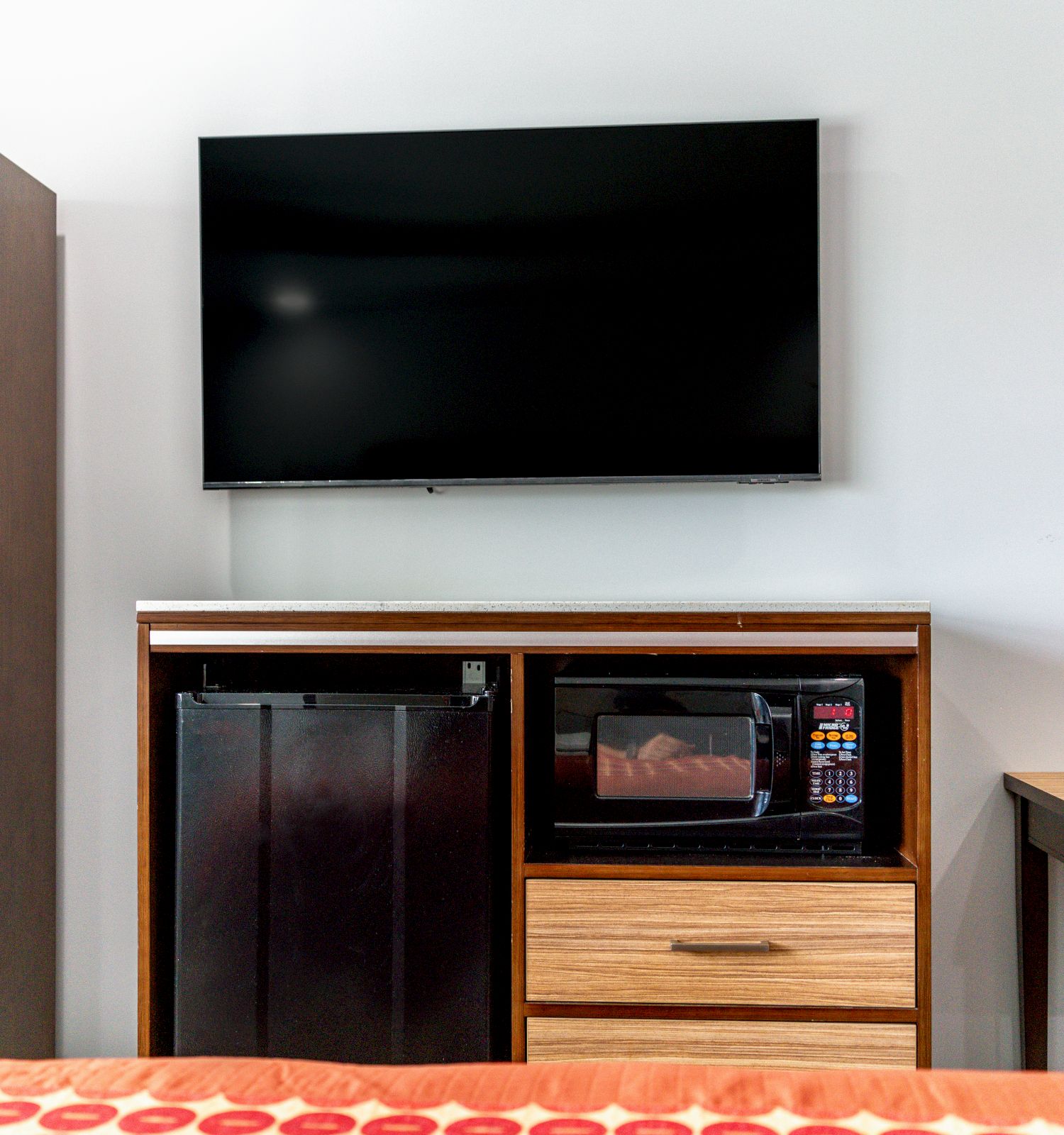 A hotel room setup with a wall-mounted TV, a cabinet housing a mini-fridge, microwave, and drawer, plus a table on the side.