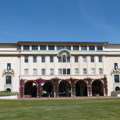 The image shows a large, white building with multiple windows and archways, surrounded by green lawns, under a clear blue sky.