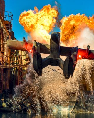 A dramatic scene of a seaplane taking off amidst a large explosion and water splash in an industrial setting, displaying intense action.