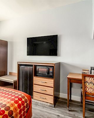 The image shows a hotel room with a bed, wardrobe, TV, mini-fridge, microwave, desk, and chair, all under a bright, fresh atmosphere.